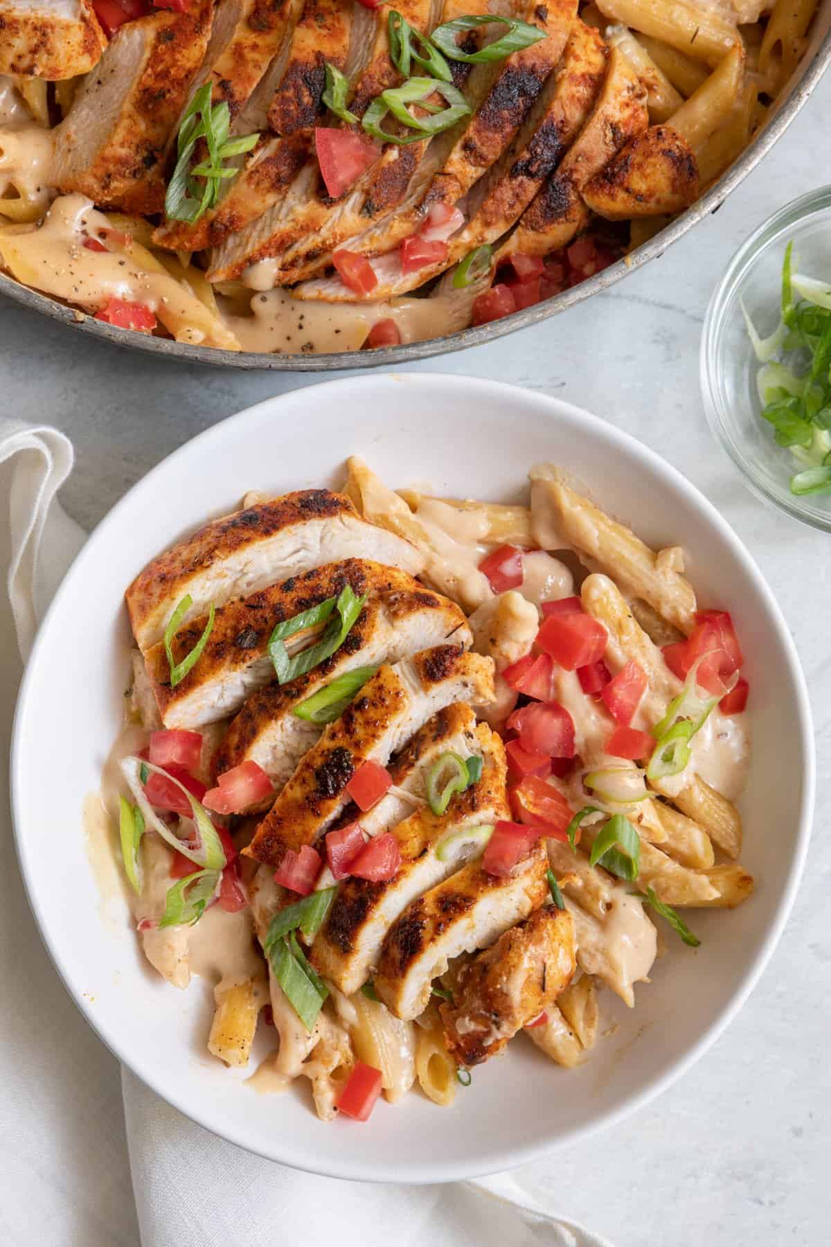 Bowl of creamy cajun pasta topped with sliced chicken breast and garnished with green onions and diced tomatoes and the skillet of recipe to the side.