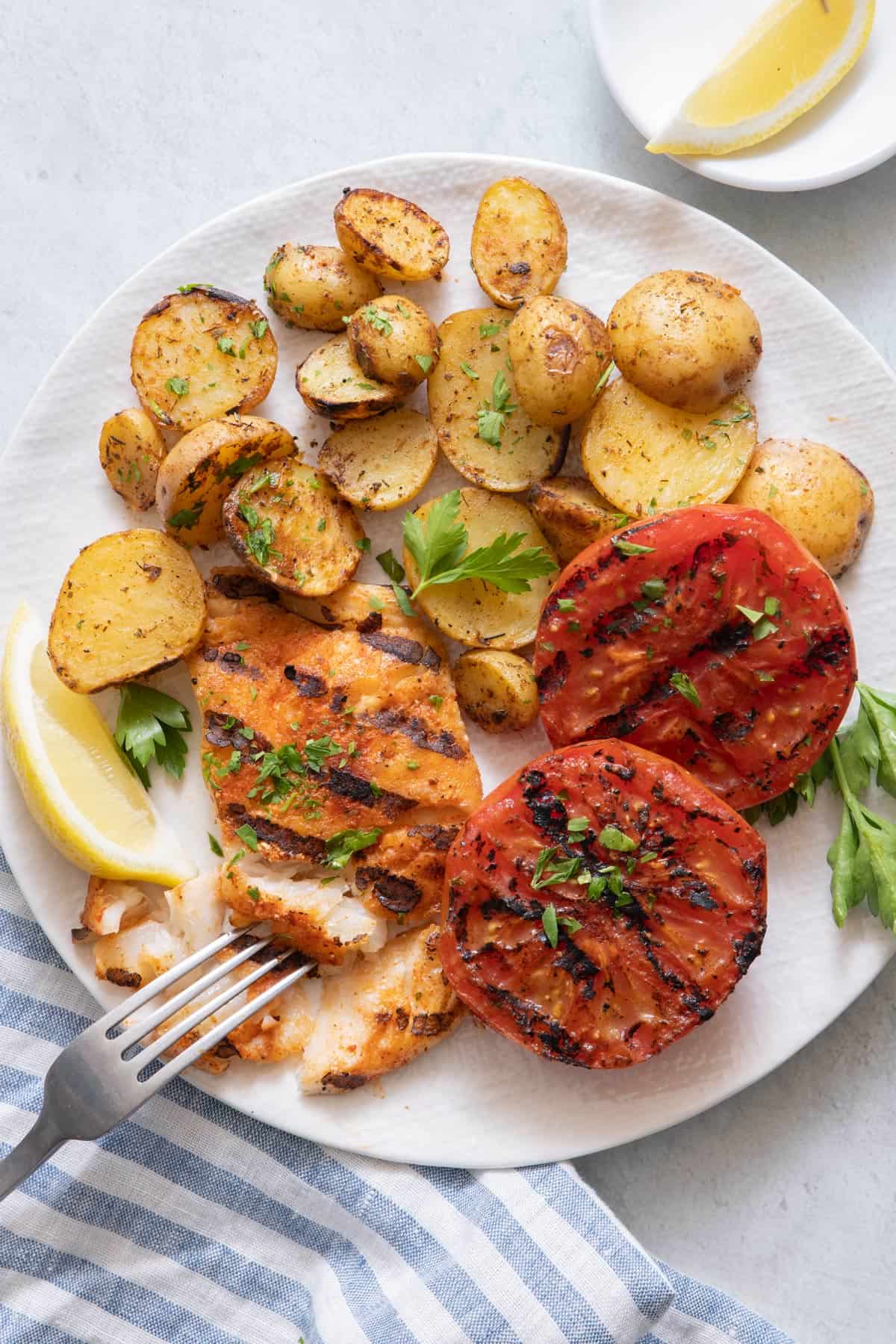 Round white plate with cod fillet with fork flaking a piece away, and plated with grilled halved baby potatoes and grilled halved tomatoes garnished with a lemon wedge and parsley..
