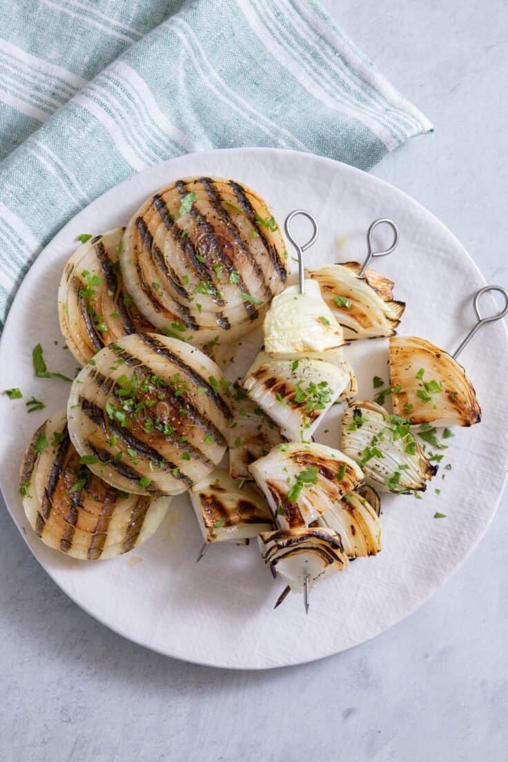 Grilled onion slices and grilled onion chunks on skewers served on a white plate with parley garnish.