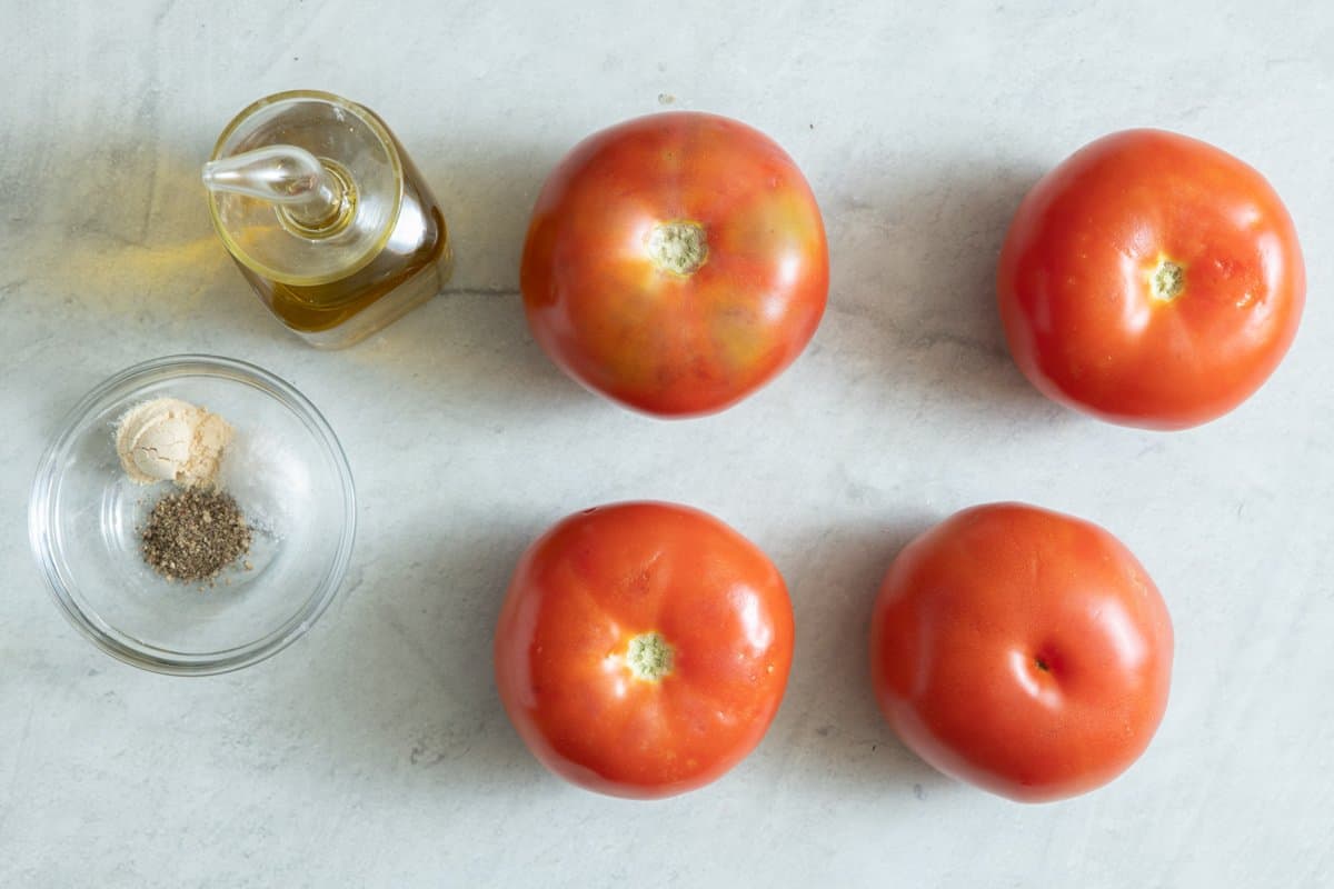 Overhead shot of 4 tomatoes, oil in a glass bottle, and a small bowl with salt, pepper, and garlic powder.
