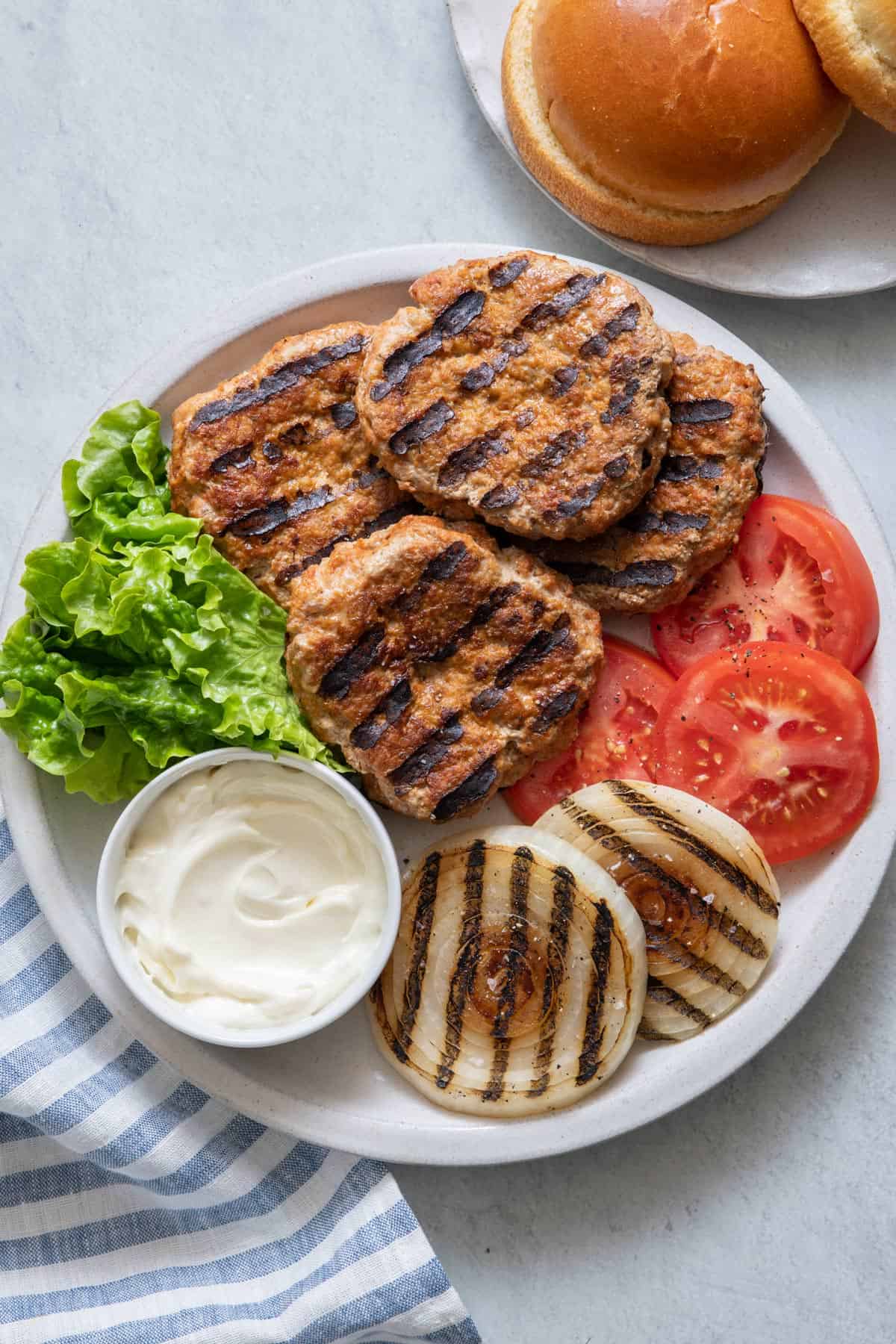 Grilled turkey burgers on plate with lettuce, tomato, grilled onions, and a small bowl of mayo with buns off to side.