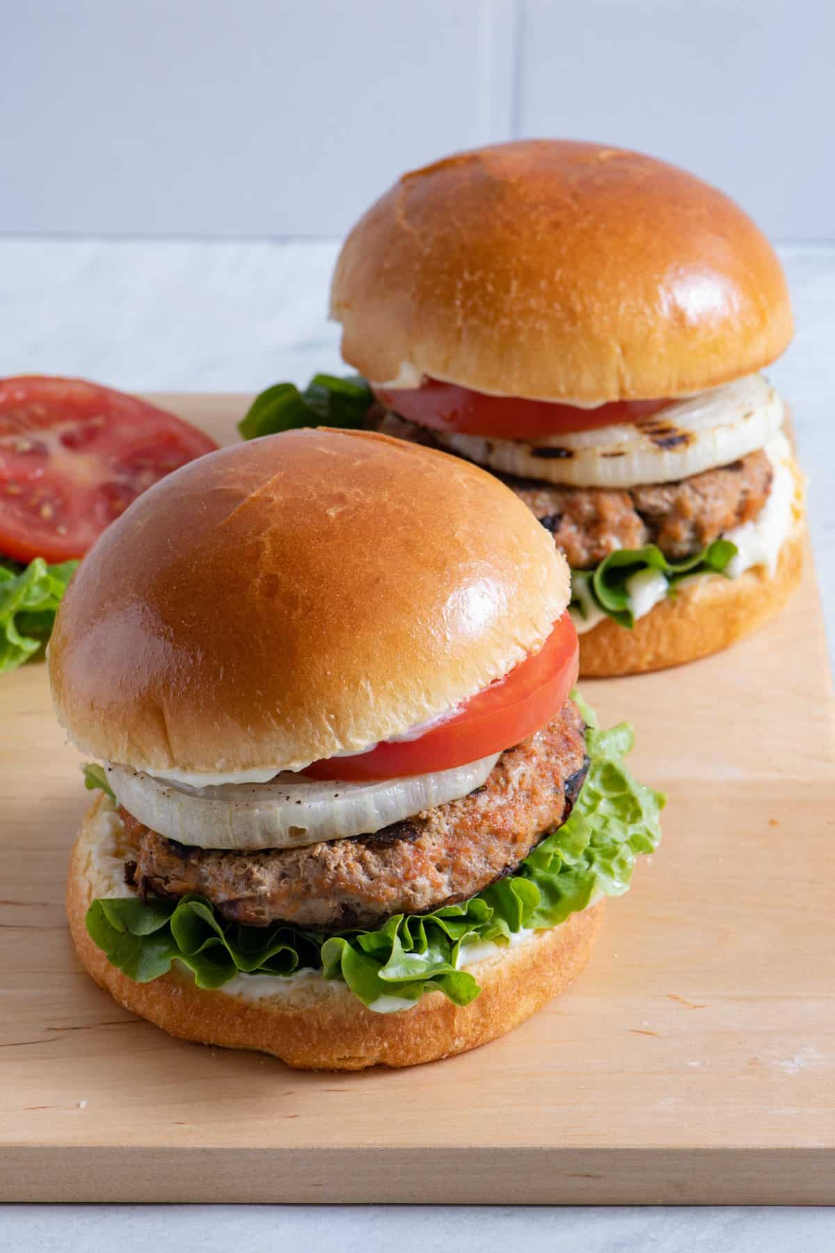 Two turkey burgers on board with lettuce, tomato, grilled onions, and mayo on bun.