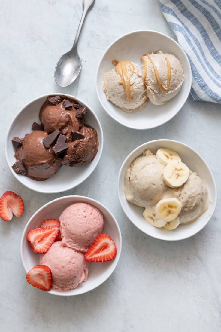 4 Nice cream flavors in white bowls: chocolate with chocolate chunks, strawberry with fresh strawberries on top, vanilla banana with banana slices on top, and peanut butter nice cream with a drizzle of peanut butter on top.