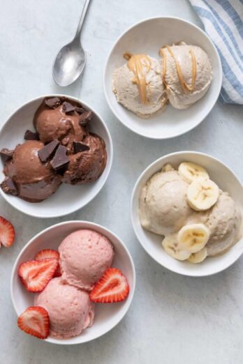 4 Nice cream flavors in white bowls: chocolate with chocolate chunks, strawberry with fresh strawberries on top, vanilla banana with banana slices on top, and peanut butter nice cream with a drizzle of peanut butter on top.