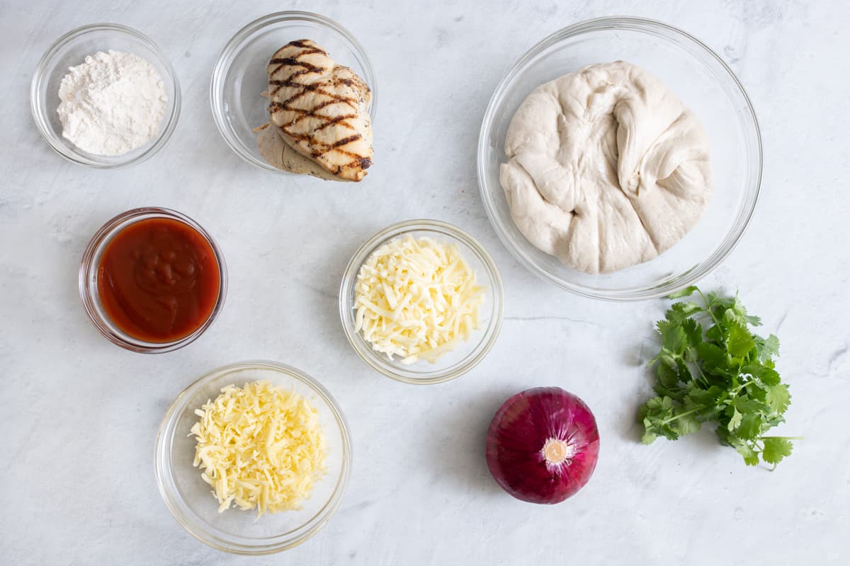 Ingredients to make pizza in individual serving bowls: flour, grilled chicken breast, bbq sauce, two bowls of different cheeses, pizza dough, whole red onion, and cilantro.