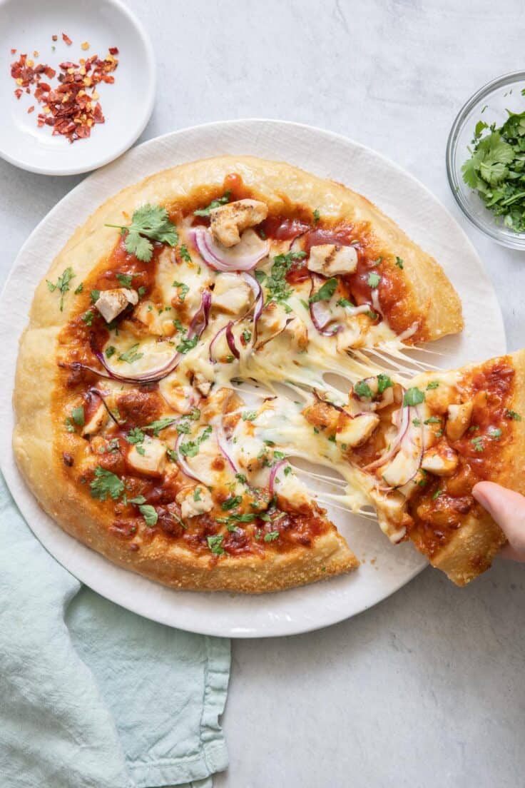 BBQ chicken pizza with a slice cut out being picked up from plate and showing melty cheese pulled from pizza.