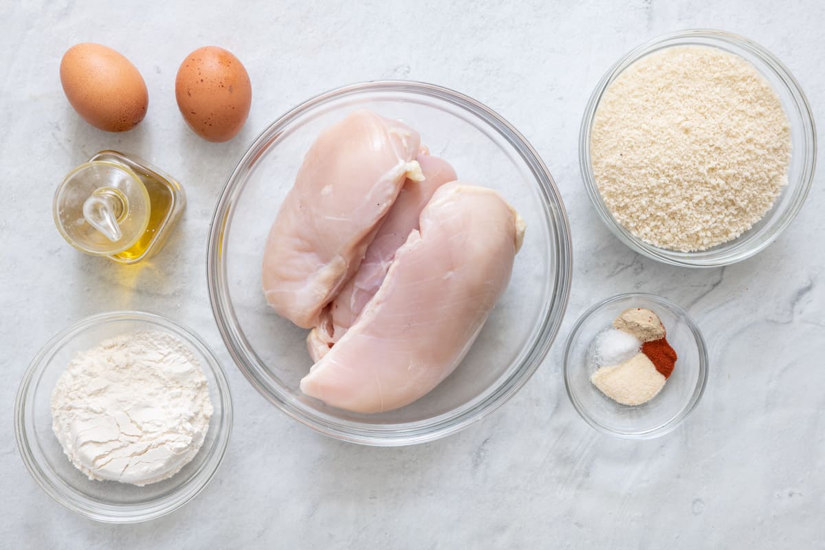 Ingredients for recipe before being prepared in and individual bowls: 2 eggs, oil, flour, chicken breast, breadcrumbs, and seasoning.