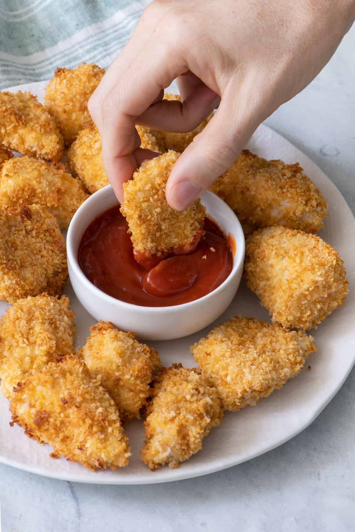 Baked chicken nugget being dunked into a small bowl of ketchup in the middle of a plate full of more chicken.