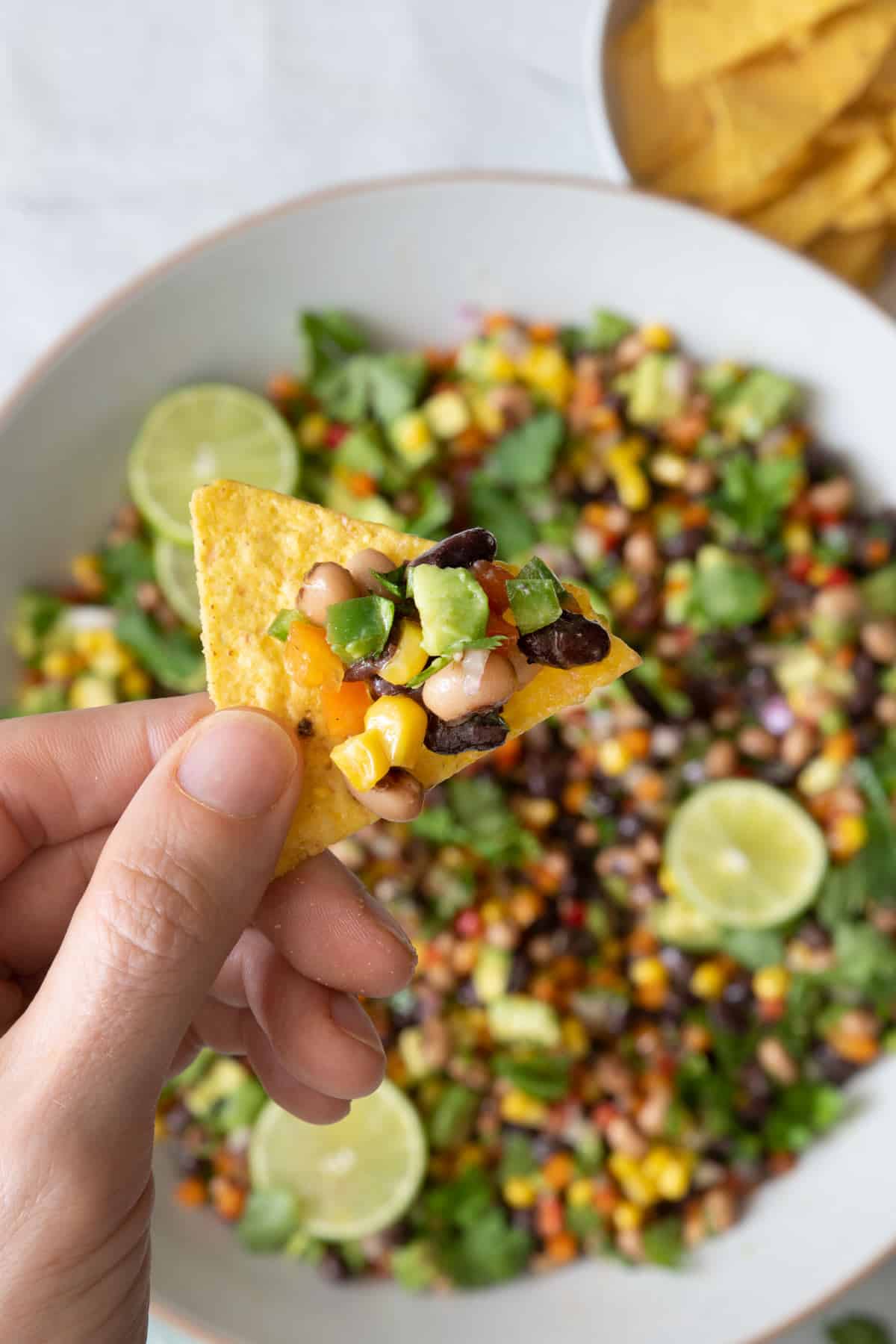 Overhead shot showing bowl of cowboy caviar underneath a hand holding a tortilla chip with recipe loaded on it.