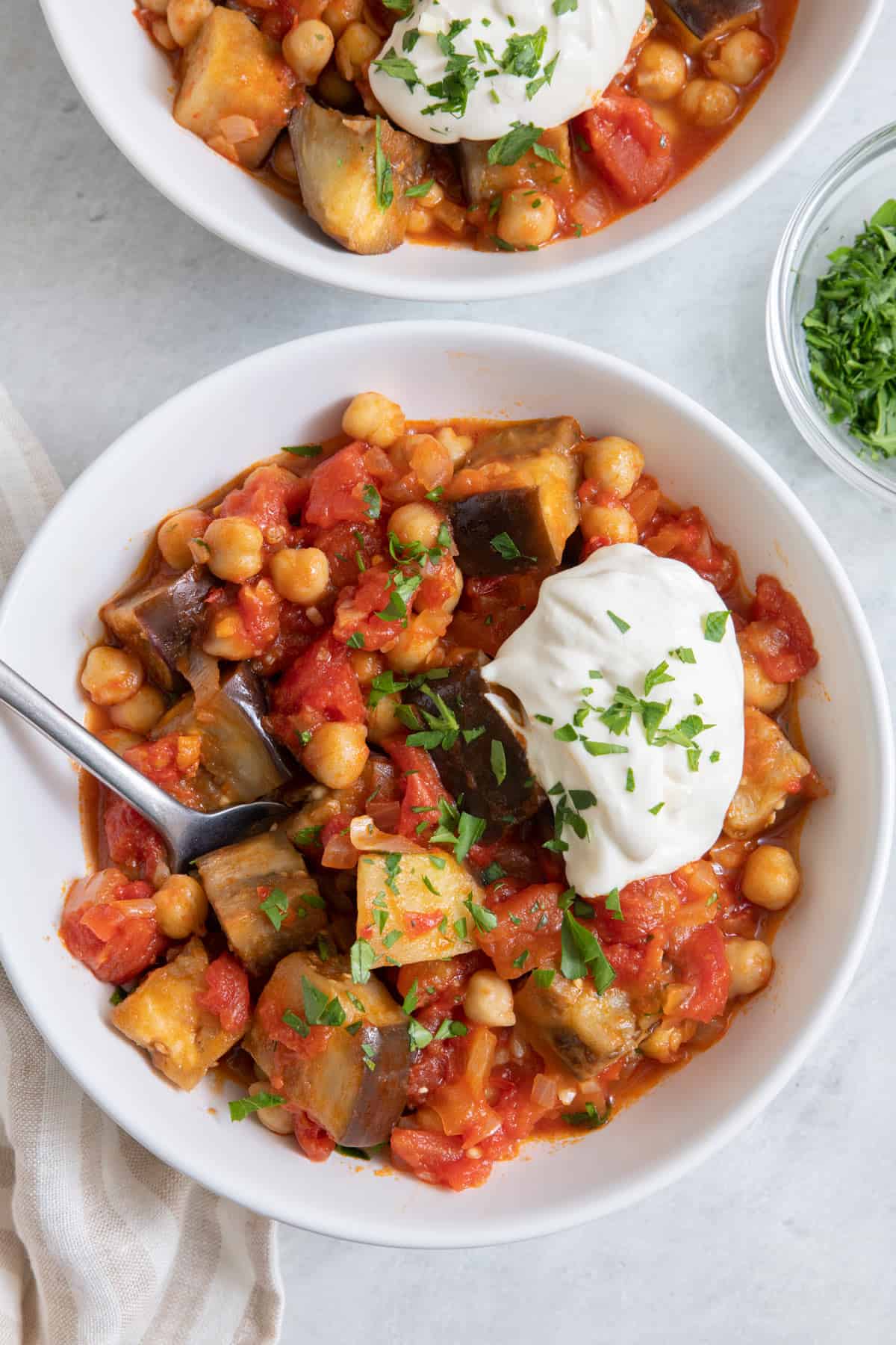 Eggplant stew served in two bowls with dollop of yogurt and parsley and spoon in one bowl, closer on one bowl.
