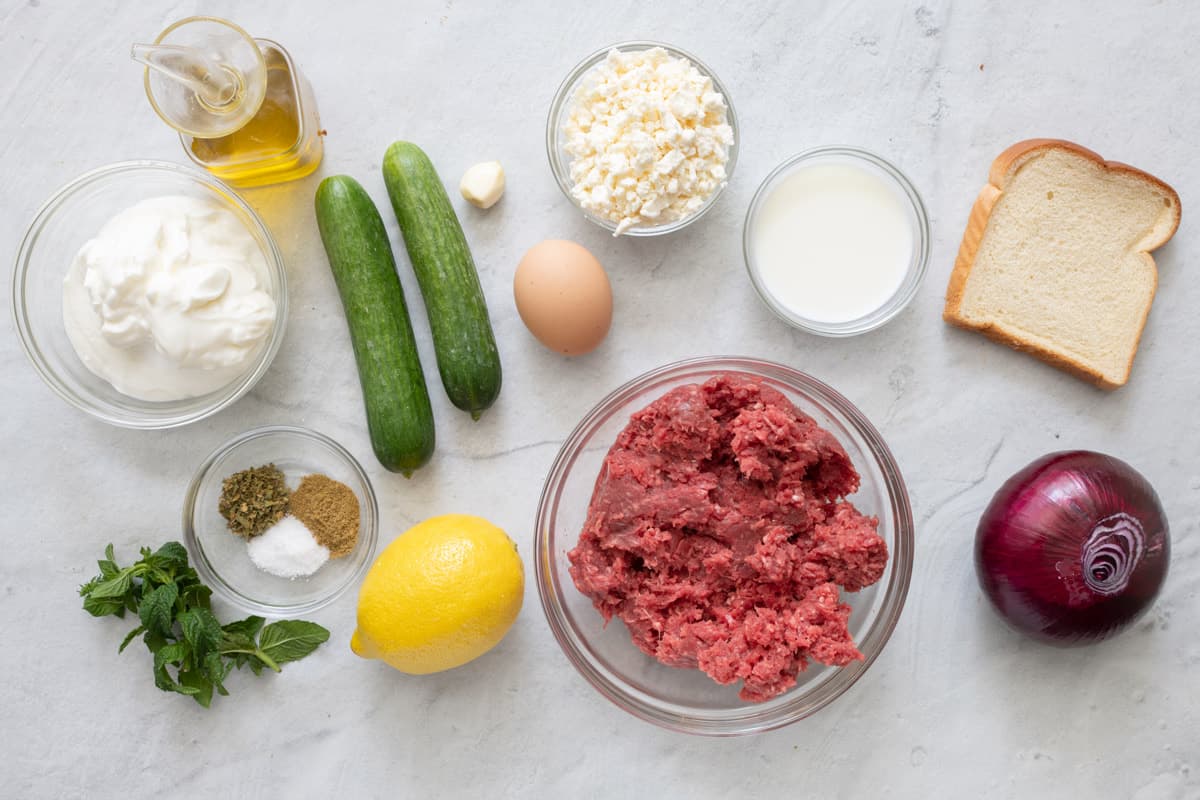 Ingredients for recipe before prepped: oil, yogurt, spices, fresh mint, 2 persian cucumbers, whole lemon, garlic clove, 1 egg, feta cheese crumbles, milk, ground beef, slice of bread, and whole red onion.