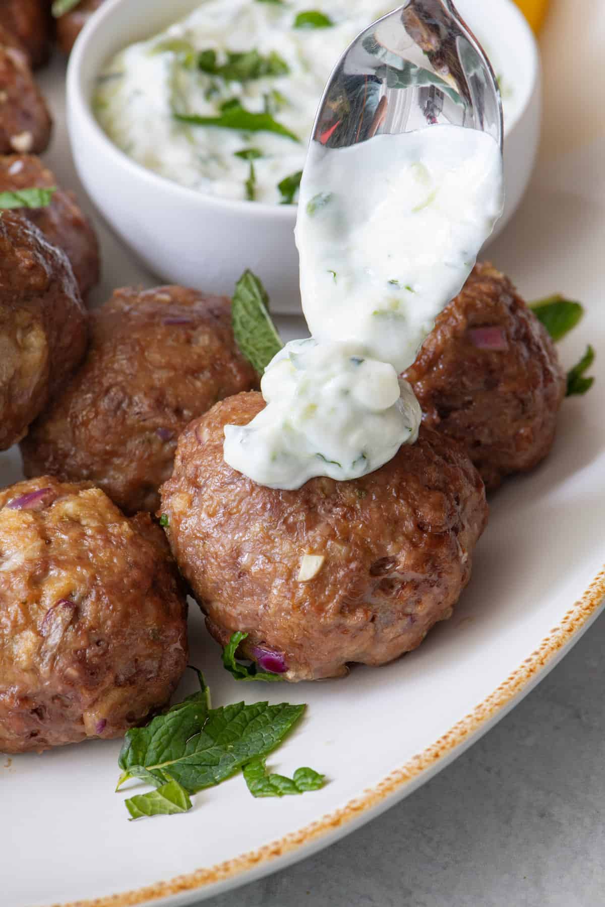 Spoon dropping tzatzki over a single meatball on a platter with more meatballs.