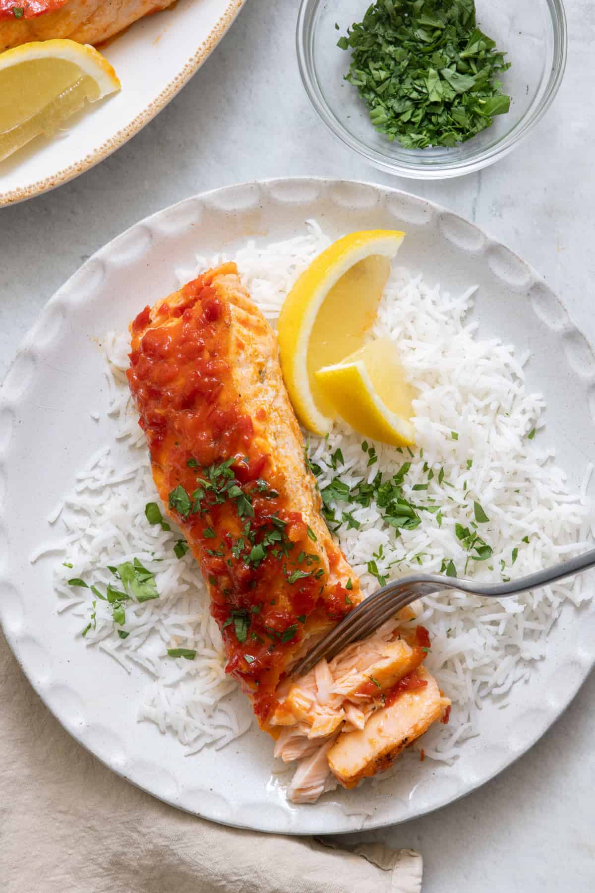 Salmon being flaked with fork over rice on a white plate with lemon wedges and parsley.