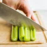 Celery stalks being cut into thirds on a cutting board with large knife.