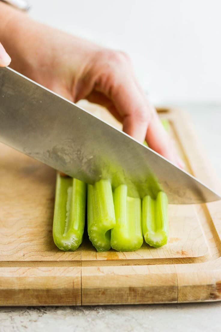 Celery stalks being cut into thirds on a cutting board with large knife.