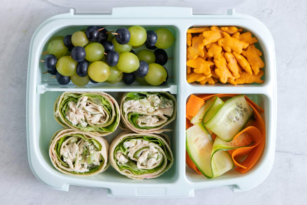 lunchbox with individual sections with different lunch foods for kids: grapes and blueberries, cheese crackers, carrot and cucumber ribbons, and chicken salad pinwheels.