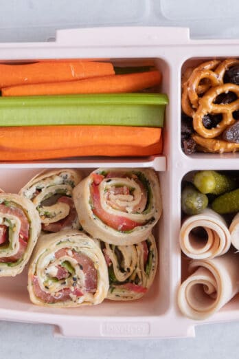 Lunchbox with individual sections with different lunch foods for kids: celery and carrot sticks, pretzels with raisins, turkey roll ups, and everything bagel pinwheels.