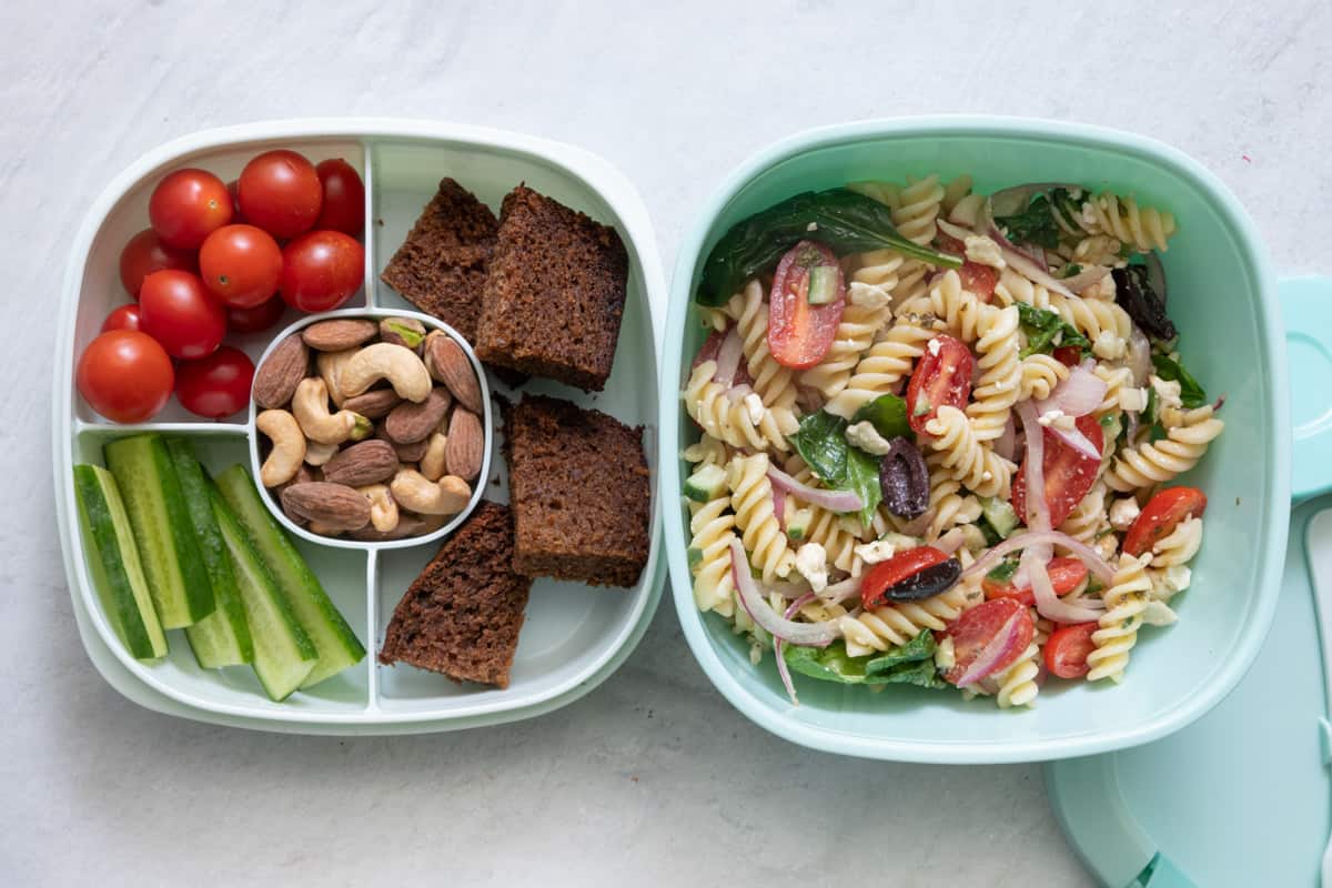 Stackable lunch container with individual sections and different foods in each: cherry tomatoes, cucumber slices, mixed nuts, apple bread slices in one container with Mediterranean pasta salad in another large container.