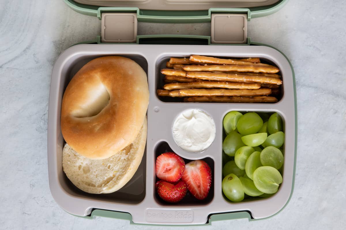Lunchbox with individual sections with different lunch foods for kids: sliced bagel, pretzel sticks, cream cheese, halved strawberries, and sliced grapes.