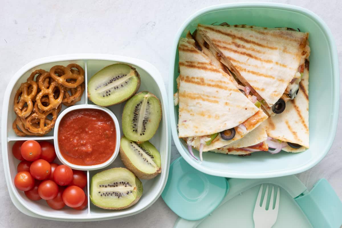 Stackable lunch container with individual sections and different foods in each: pretzels, cherry tomatoes, pizza sauce, 2 kiwis cut in half, with pizzadillas in large container.