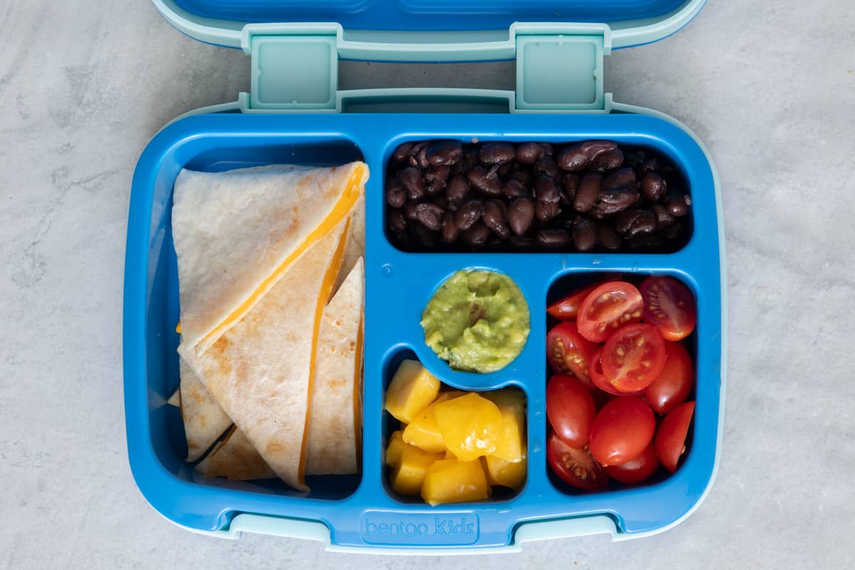 Bento blue lunchbox with individual individual sections with different lunch foods for kids: cheese quesadilla, black beans, guacomole, diced peaches, and cherry tomatoes sliced in half.