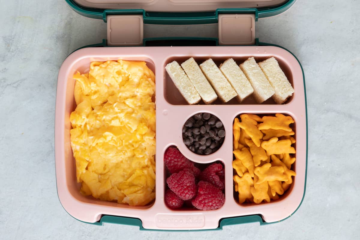 Lunchbox with individual sections with different lunch foods for kids: scrambled eggs, 6 square cut white bread, chocolate chips, raspberries, and Annie's bunnies.