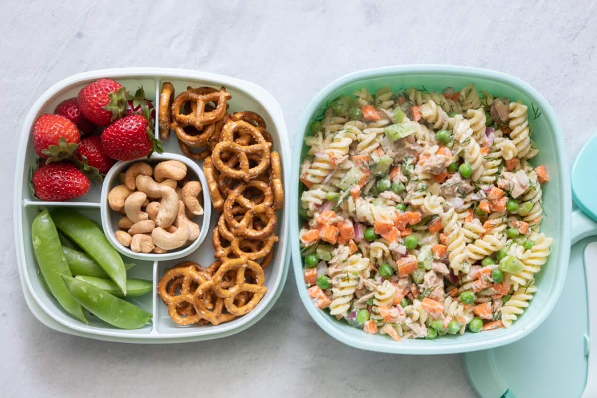 Stackable lunch container with individual sections and different foods in each: strawberries, sugar snap peas, cashews, and pretzels, with tuna pasta salad in large container.