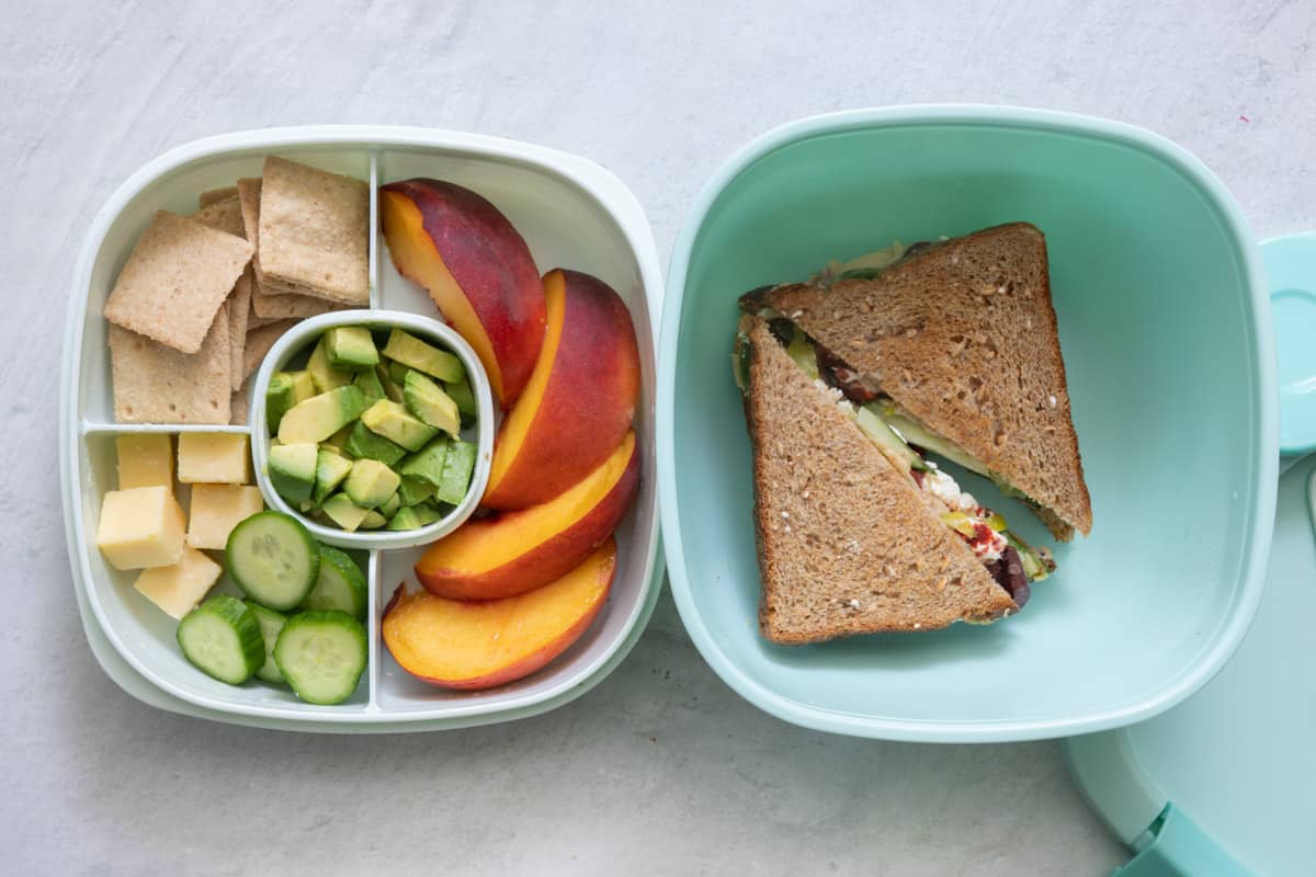 Stackable lunch container with individual sections and different foods in each: croackers, cheese cubes with cucumbers, diced avocado, and sliced peaches in one container with a veggie hummus sandwich in the other.