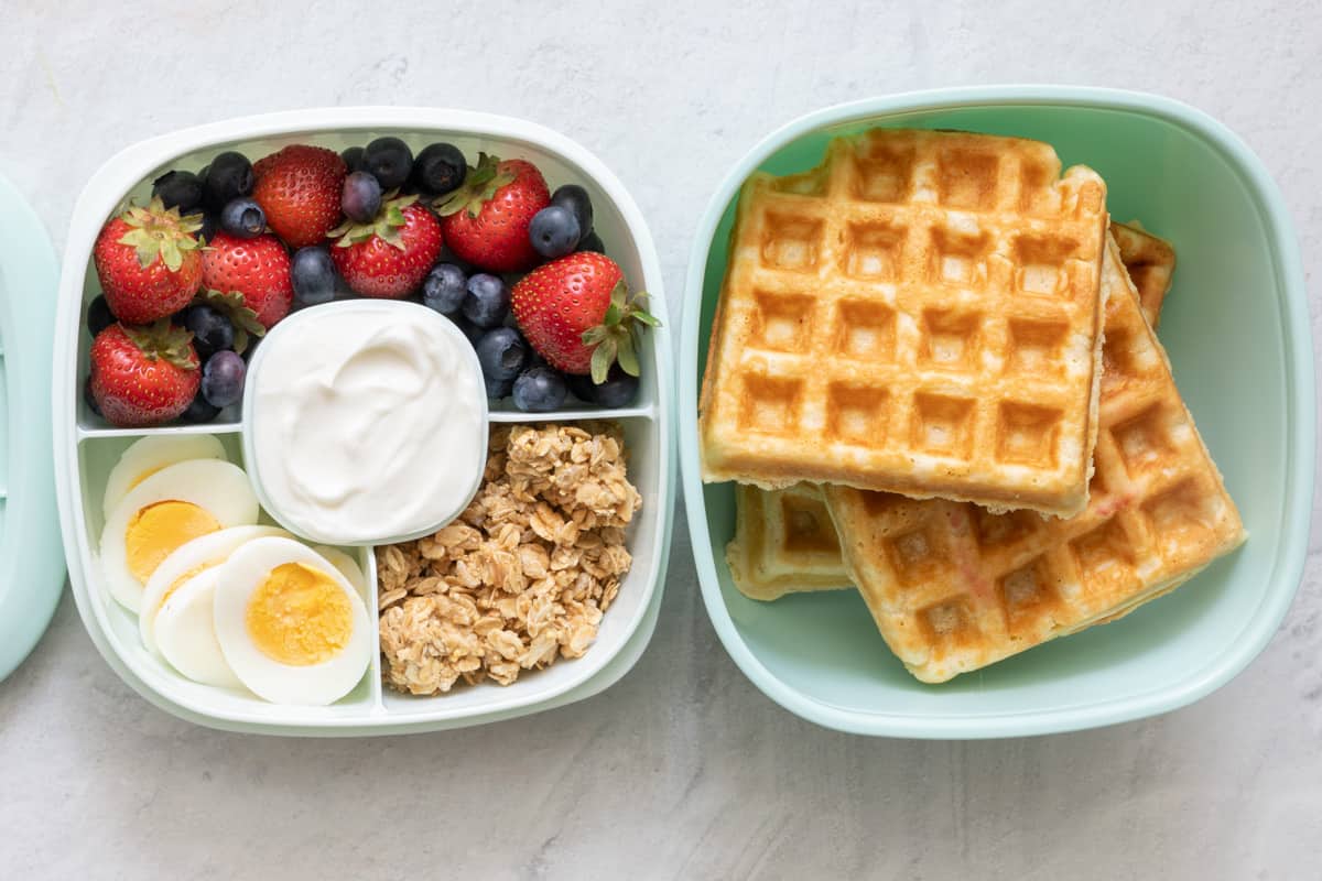 Stackable lunch container with individual sections and different foods in each: strawberry and blueberries, sliced boiled egg, greek yogurt, and granola with 3 waffles.