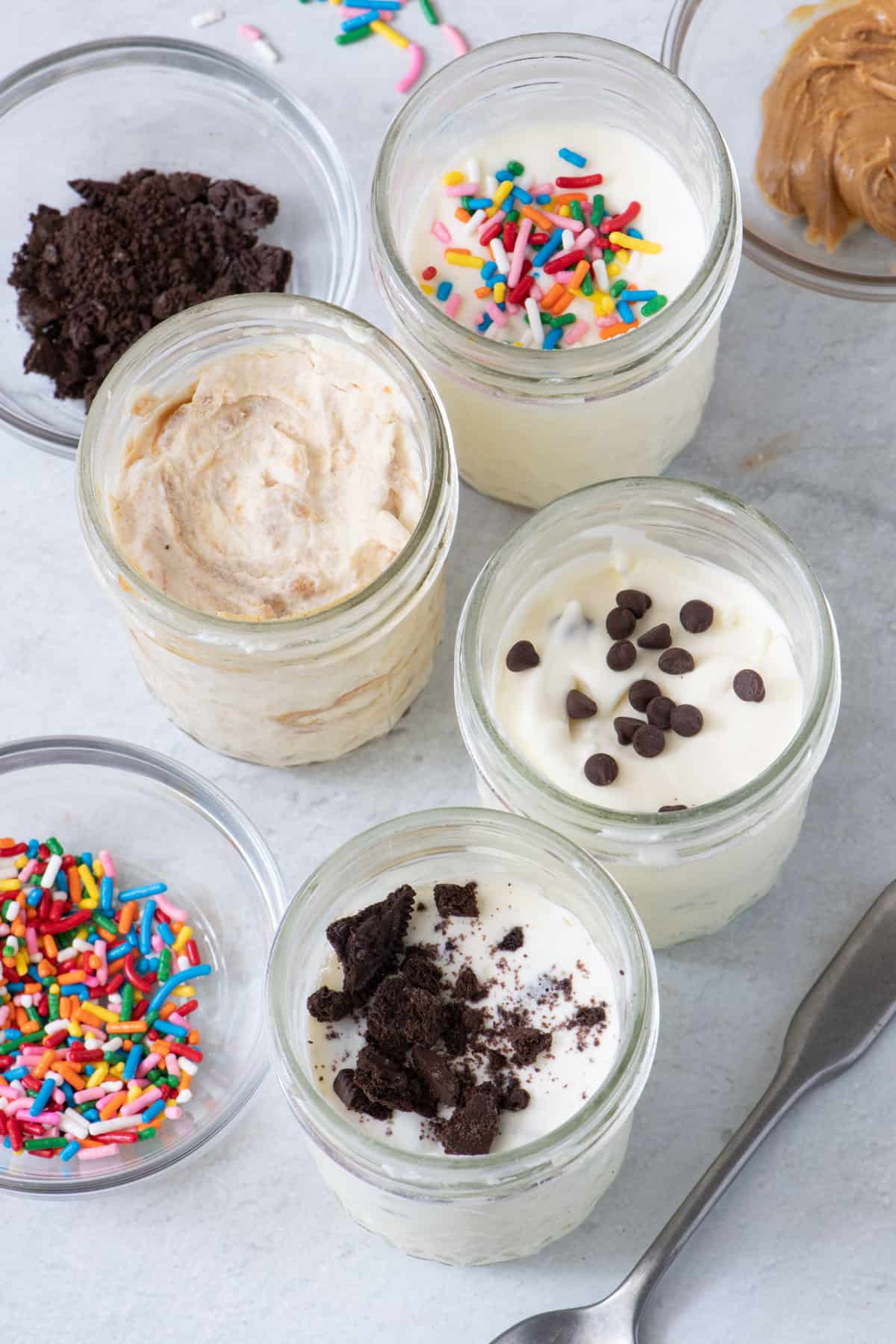 Angle view of 4 masons jars filled with ice cream and each with different toppings inside and in individual bowls placed around jars.