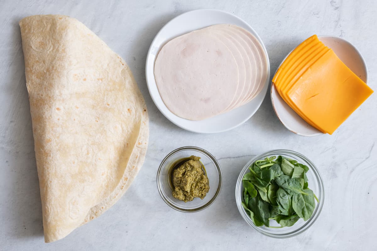 Ingredients for recipe: large tortilla shell, sliced turkey, sliced cheddar cheese, pesto, and spinach.