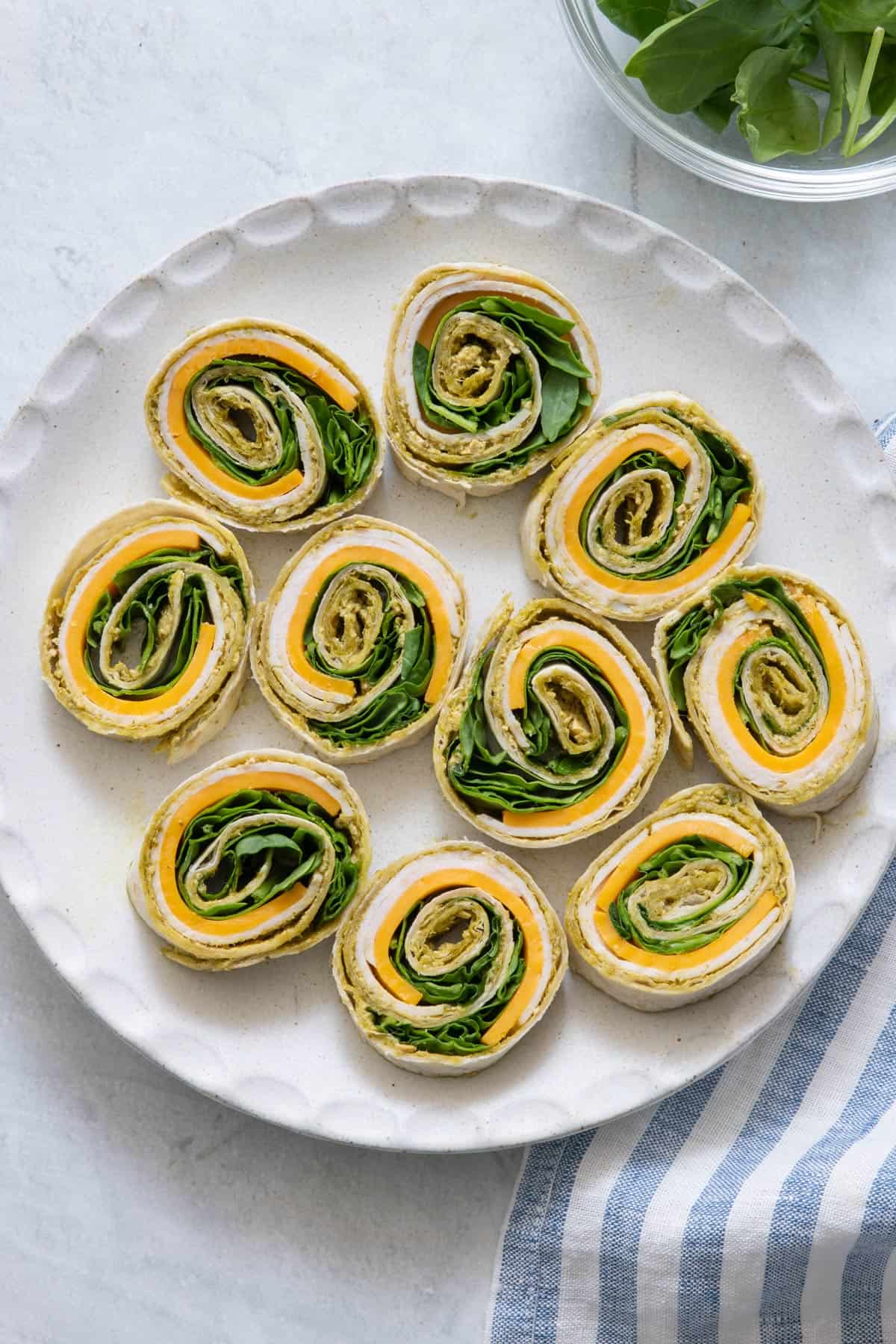 Pesto turkey pinwheel sandwiches on a round plate with bowl of fresh spinach off screen on sides.