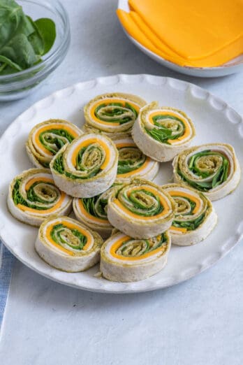 Pinwheel sandwich with spinach, turkey, cheddar cheese, and spinach, stacked on a round plate with extra ingredients in background.