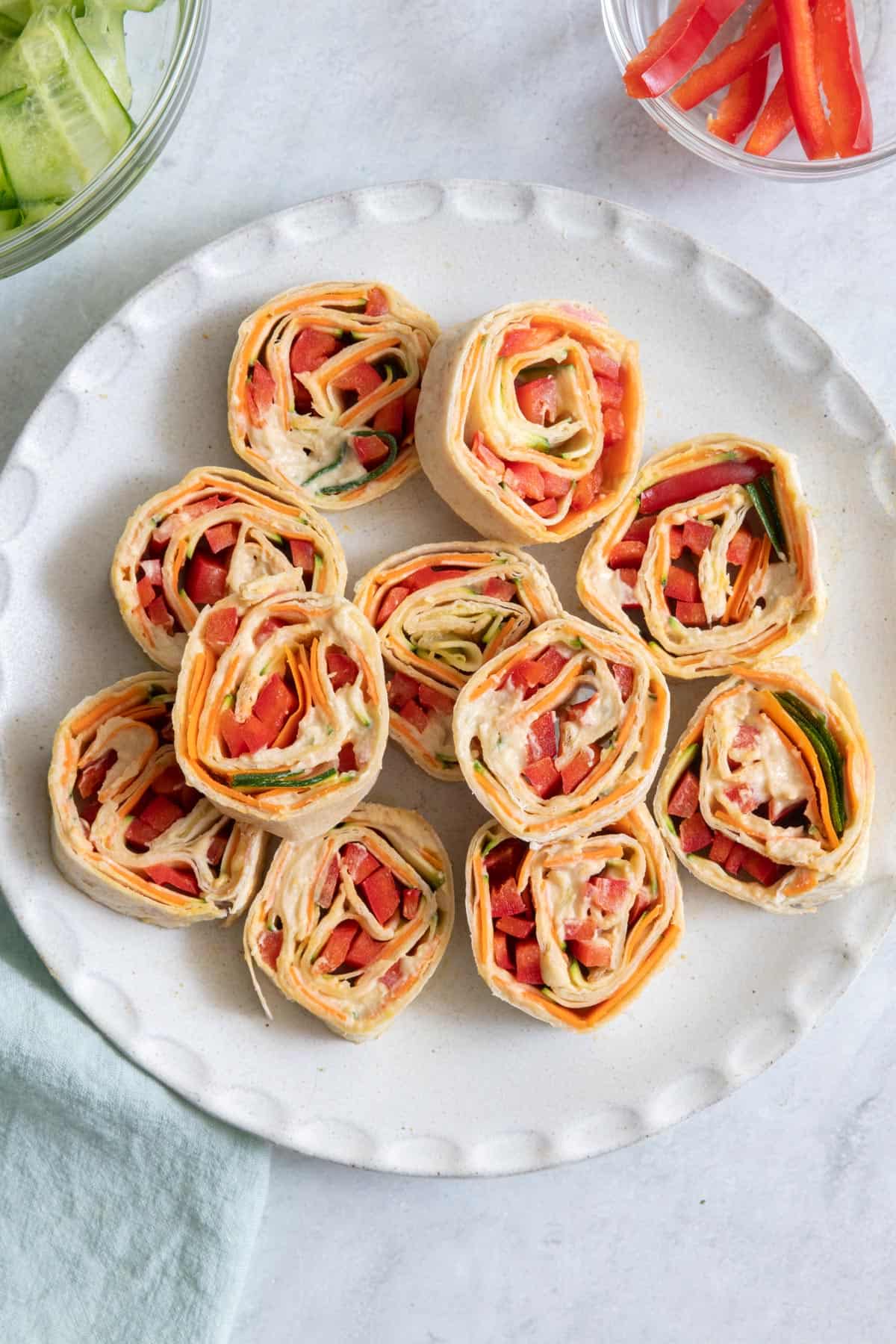Hummus and veggie pinwheel sandwiches on a round plate with ingredients off screen on sides.