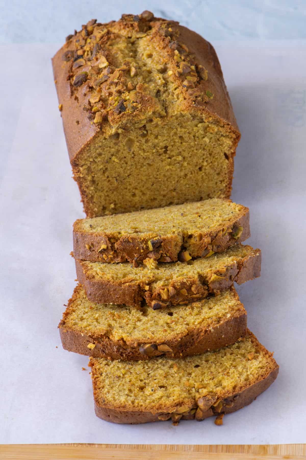 Loaf of sweet bread made with pistachios on a piece of parchment paper with four thick slices cut from half of the loaf.