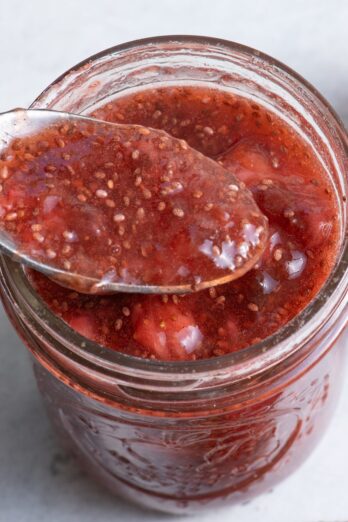 Strawberry chia jam in a jar with a spoon holding up some and strawberries nearby.