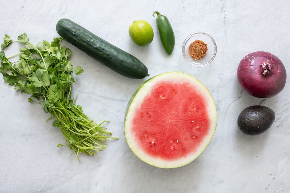 Ingredients for salsa recipe before being prepped: fresh cilantro, cucumber, lime, jalapeno, half watermelon, spices, red onion, and avocado.