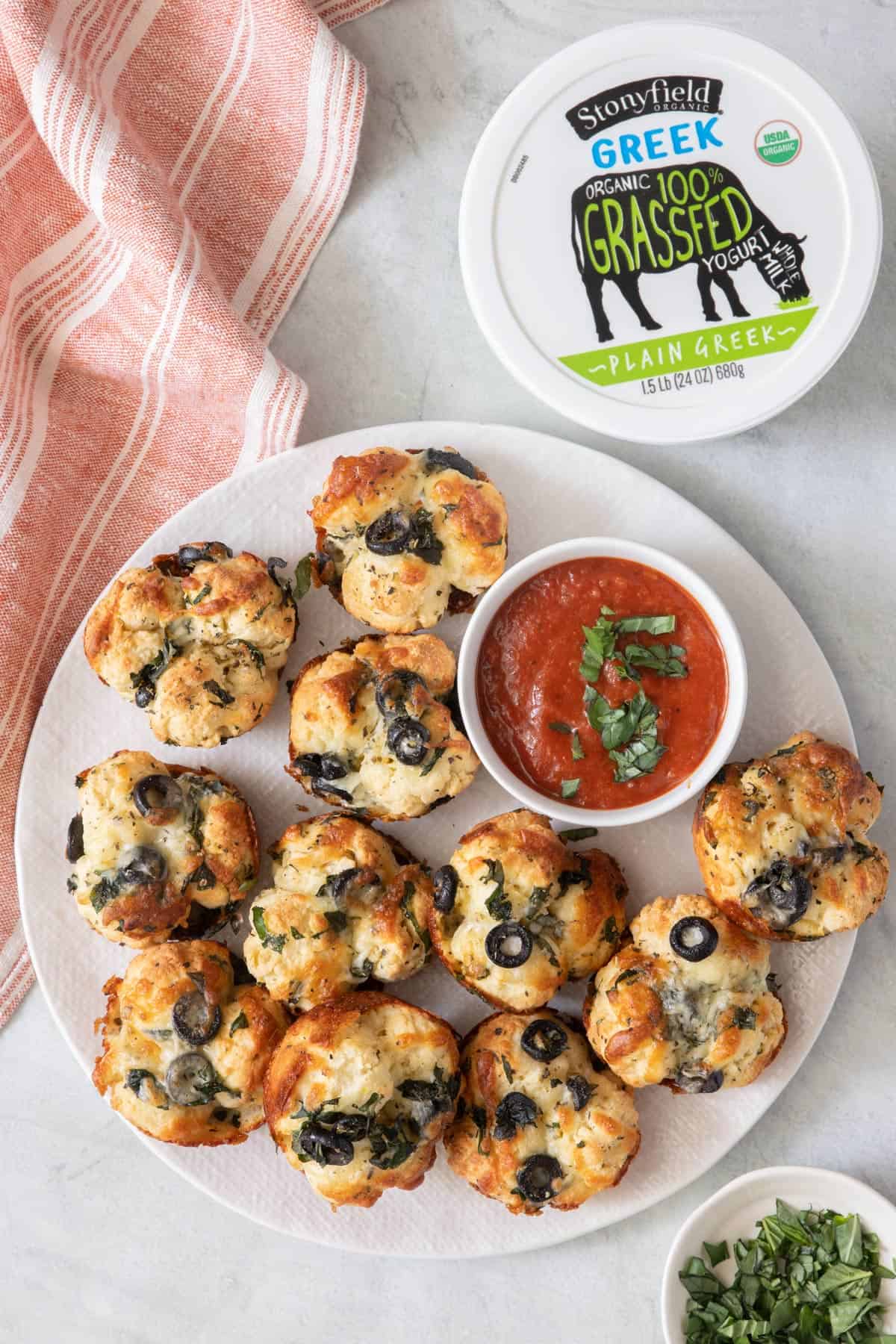 Pizza bites in round plate with pizza sauce for dipping and a tub of Greek yogurt to the side.
