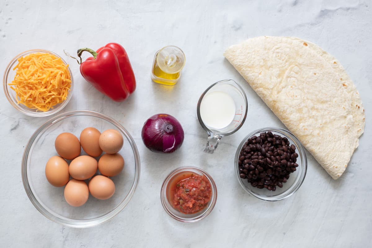 Ingredients for recipe before being prepped: shredding cheese, whole eggs, red pepper, oil, red onion, salsa, milk, black beans, and a large tortilla shell folded in half.