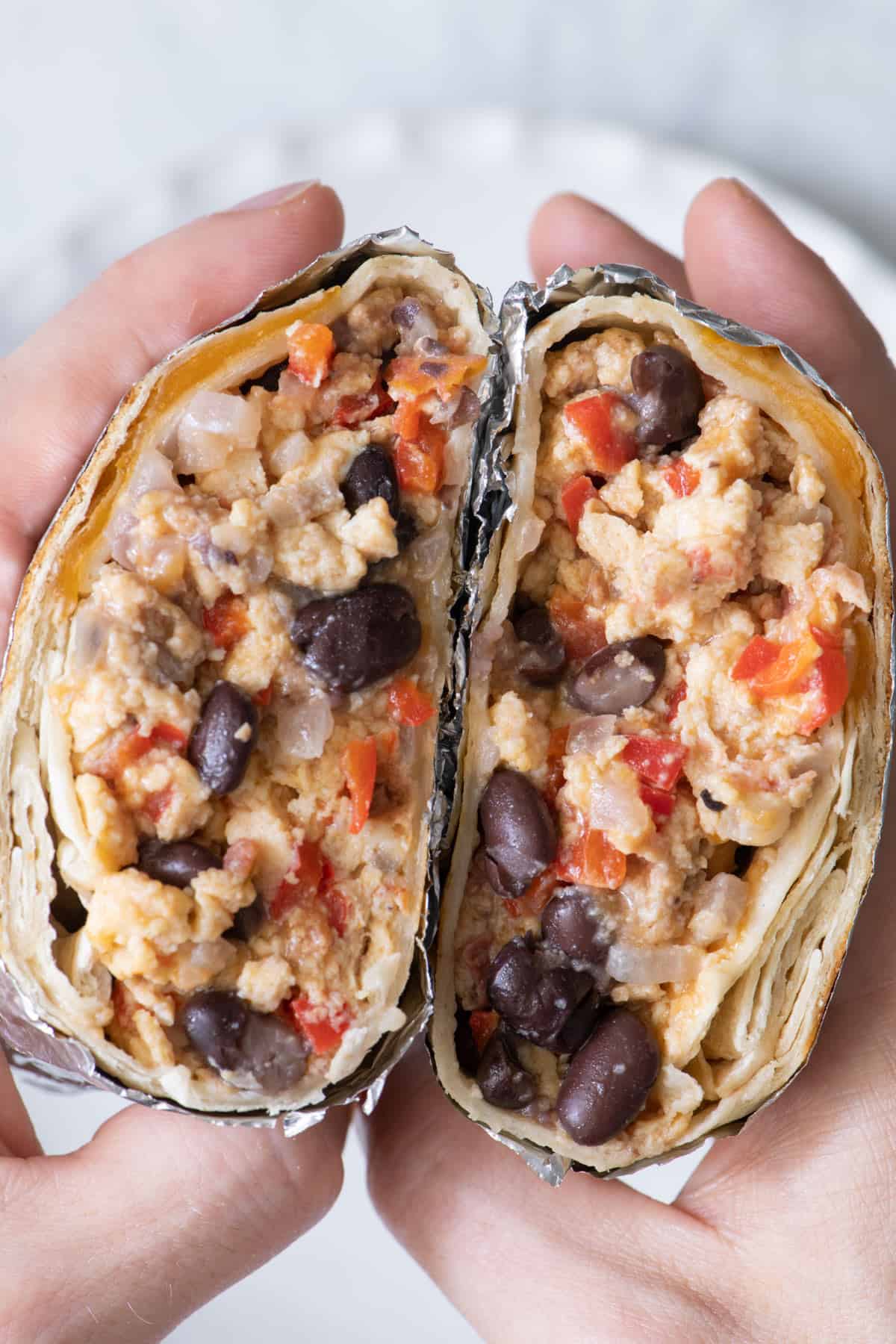 One burrito wrapped in foil, cut in half with hands holding both halves side by side.