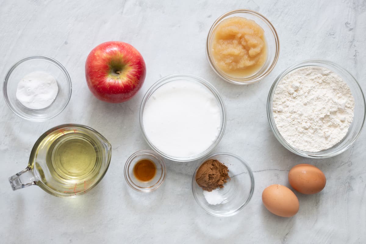 Ingredients for recipe in individual bowls: baking soda, oil, apple, vanilla, sugar, salt and cinnamon, applesauce, flour, and 2 whole eggs.