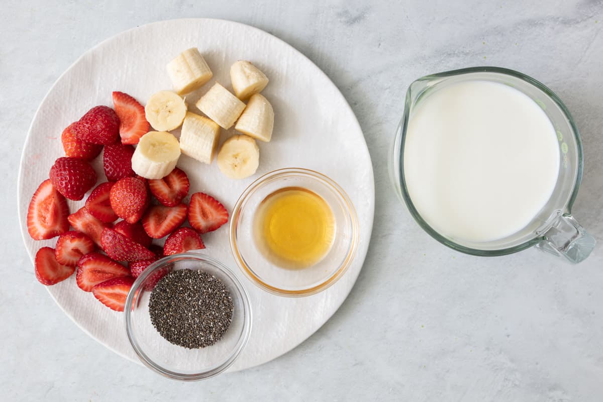Ingredients for recipe on a round plate and in individual bowls: strawberries cut in half, banana chunks, chia seeds, honey, and milk.