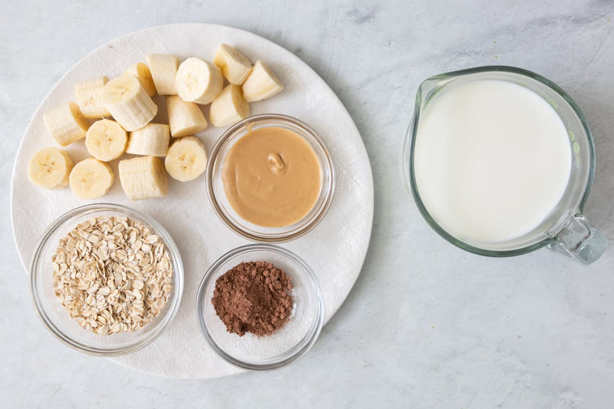 Ingredients for recipe on a round plate and in individual bowls: chunks of banana, oats, peanut butter, cocoa powder, and milk.