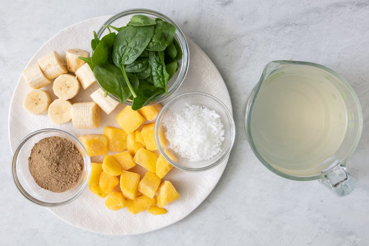 Ingredients for recipe on a round plate and in individual bowls: spinach, banana chunks, mango chunks, ground flax seed, shredded coconut, and coconut water.