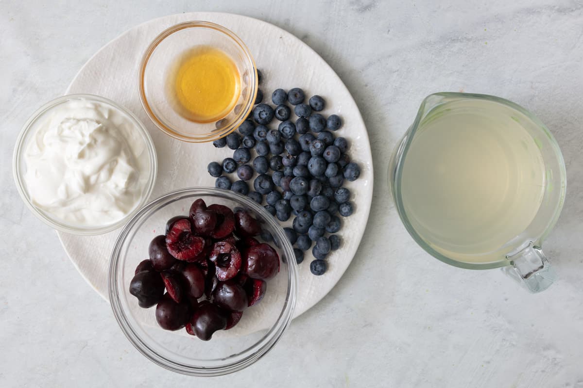 Ingredients for recipe on a round plate and in individual bowls: yogurt, honey, cherries, blueberries, and coconut water.