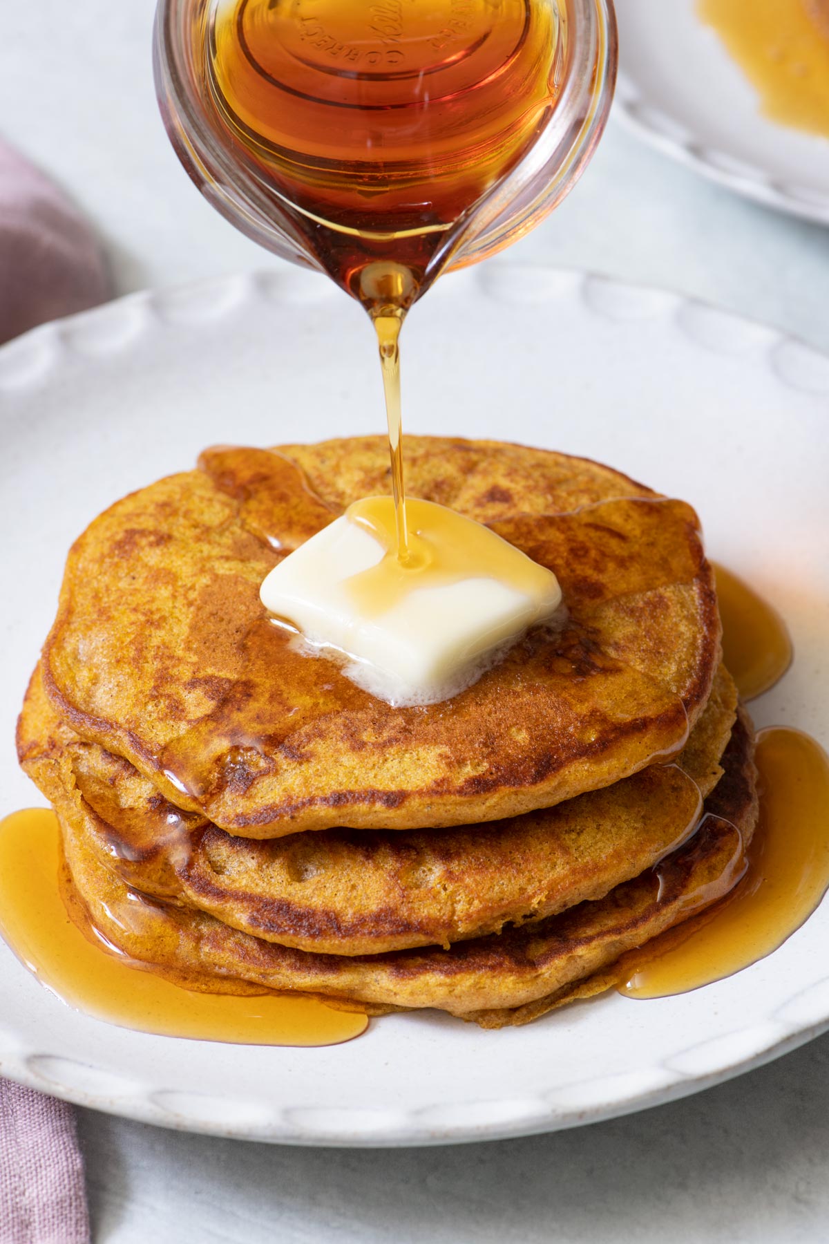 Stack of few pumpkin pancakes with dab of butter and maple syrup getting poured on.