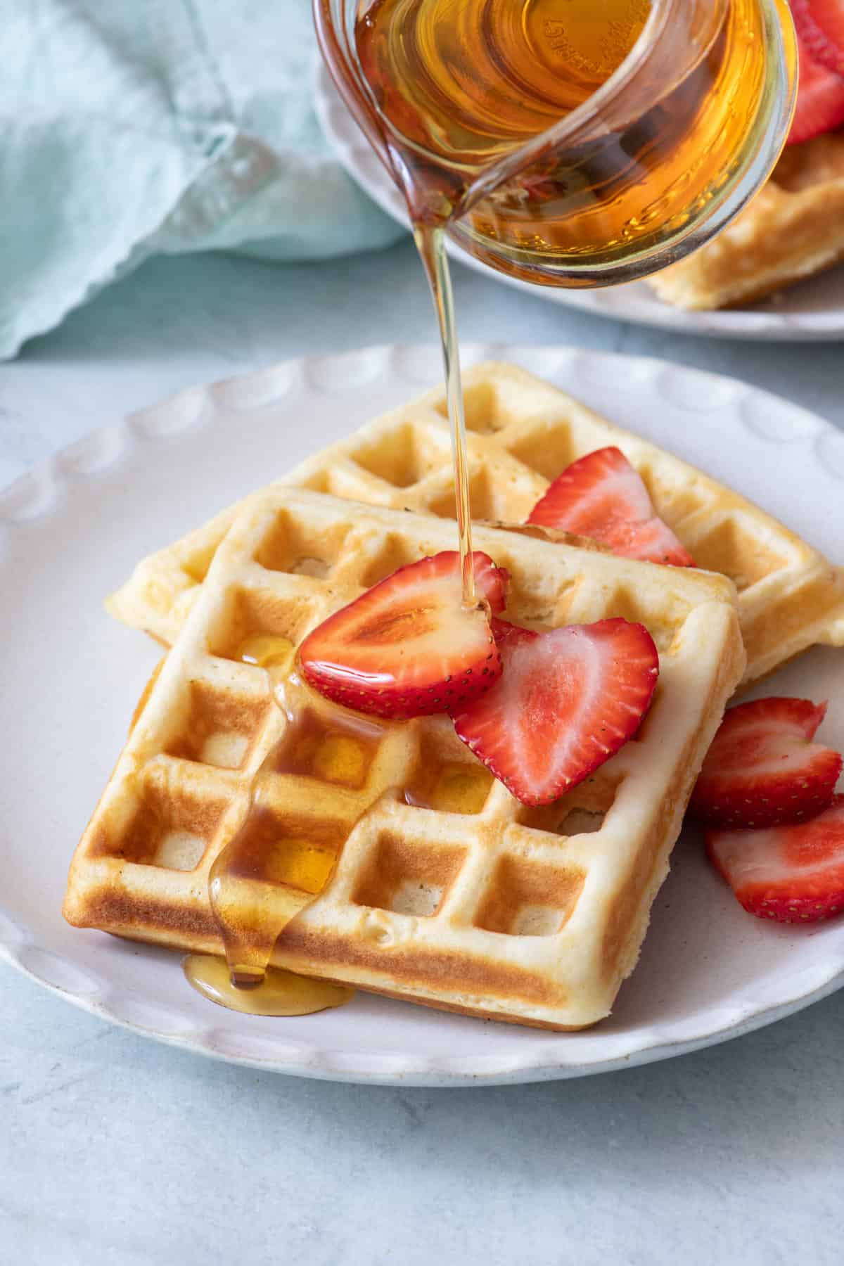 Plate of homemade waffles with slices of strawberries and maple syrup being poured on top in a glass carafe.