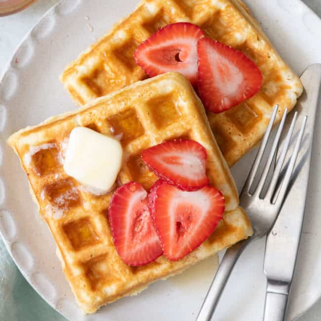 2 square waffles on a white plate with fork and knife, topped with sliced strawberries and butter.