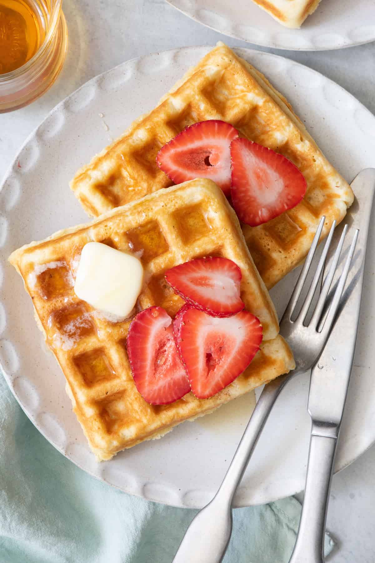 2 square waffles on a white plate with fork and knife, topped with sliced strawberries and butter.