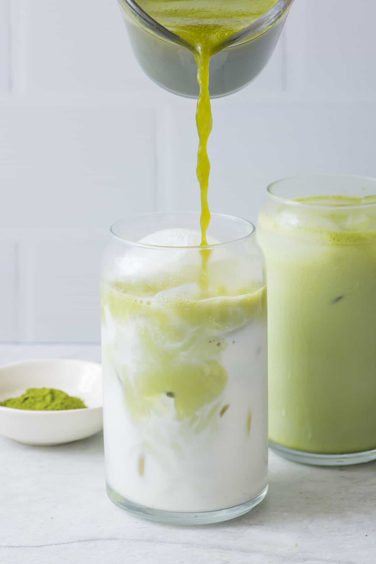 Matcha mixture being poured on top of a glass of iced milk with an additional mixed drink behind it.